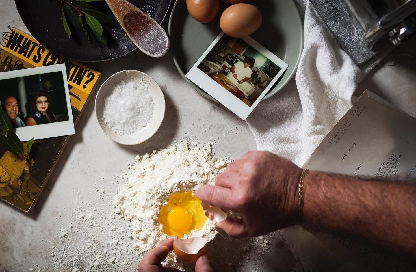 Hands cracking eggs into a flour well for fresh pasta, with polaroid photos in the background