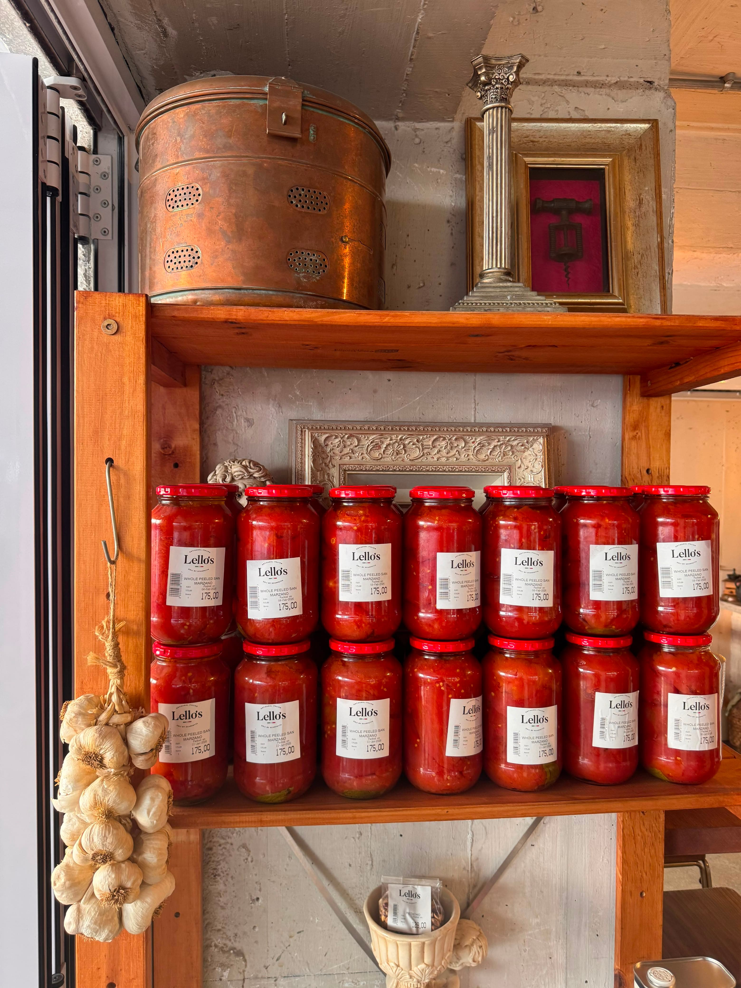 Shelf stacked with jars of Lello's homemade tomato sauce, garlic braid hanging alongside