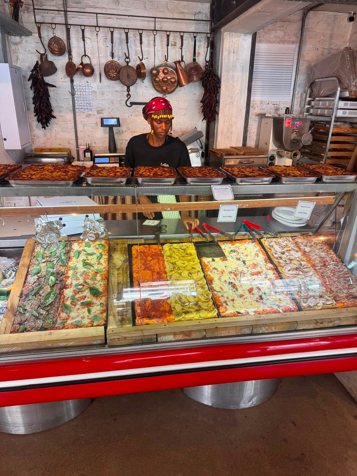 Lello's pizza al taglio display counter stacked with fresh trays and copper pans on the wall